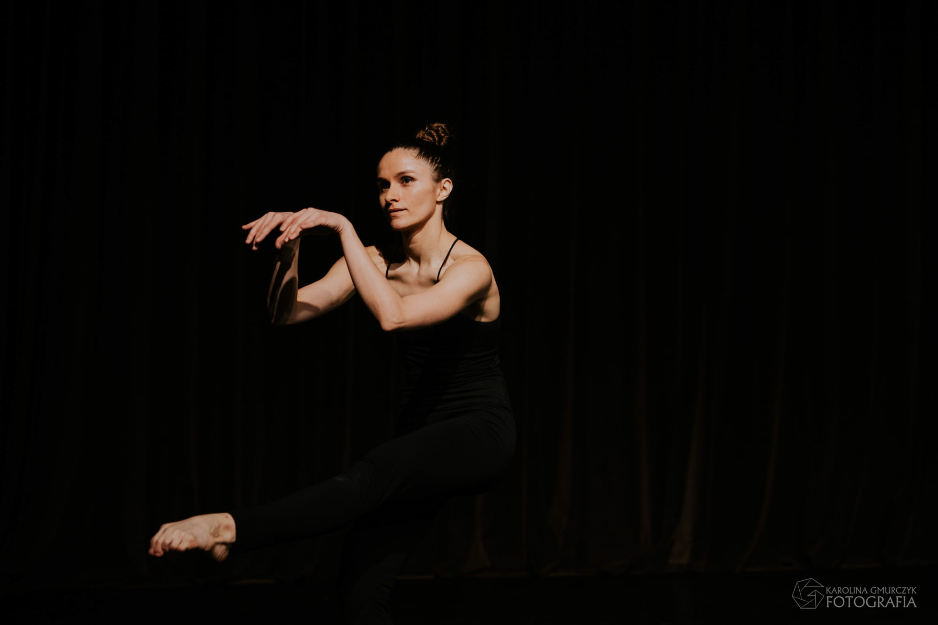 Zdjęcie, kobieta na czarnym tle wykonuje układ choreograficzny. Uśmiecha się.